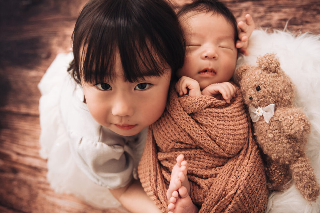 茶色のニットラップで包まれて眠る新生児に、お姉ちゃんが寄り添う家族撮影プランの作品例。バスケットの中、木目背景に毛皮のラグを敷いたあたたかな一枚。