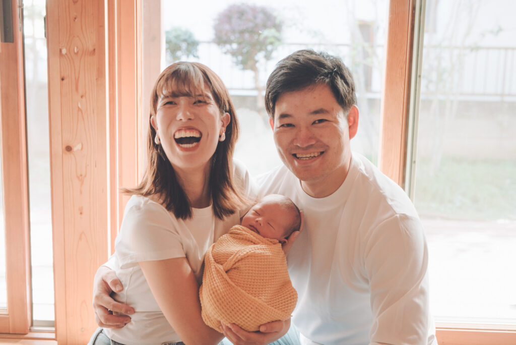 窓辺の縁側で、赤茶ニットラップの新生児を抱いて笑顔を見せる若いご夫婦のあたたかい家族写真