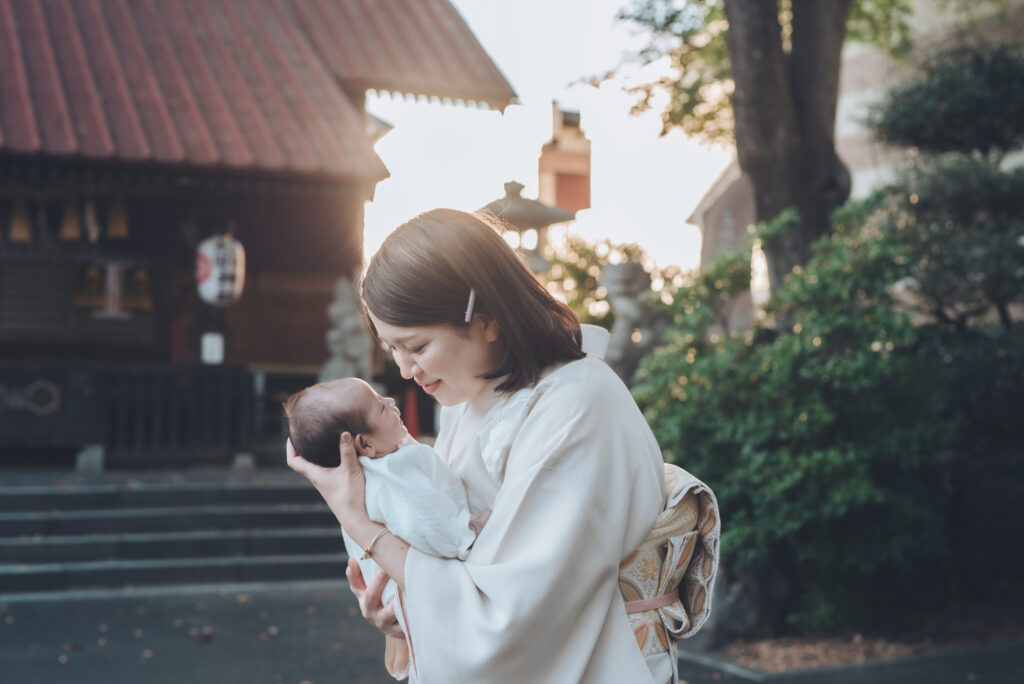 神社の境内で、白い着物姿のお母さんがやさしく赤ちゃんを抱き上げて見つめている、お宮参りの一枚。夕日の柔らかい光に包まれている。